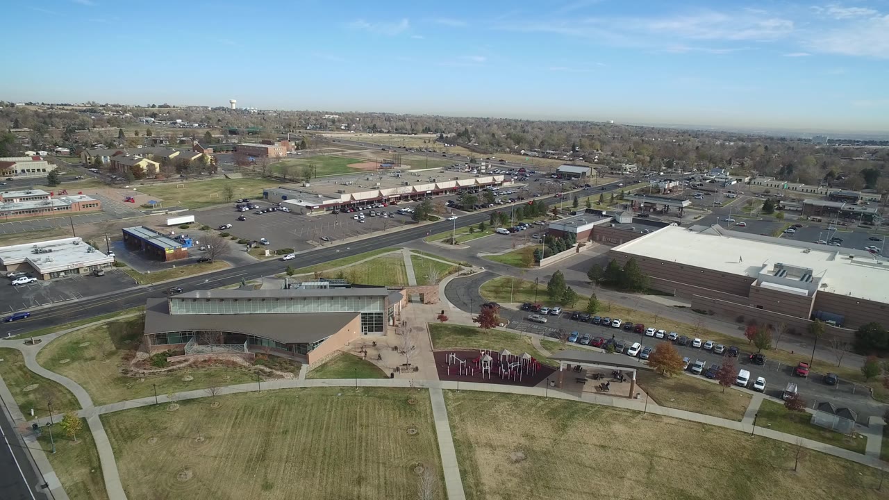 Aerial frame of a large building, lawns, roads, parked cars, and suburban development.