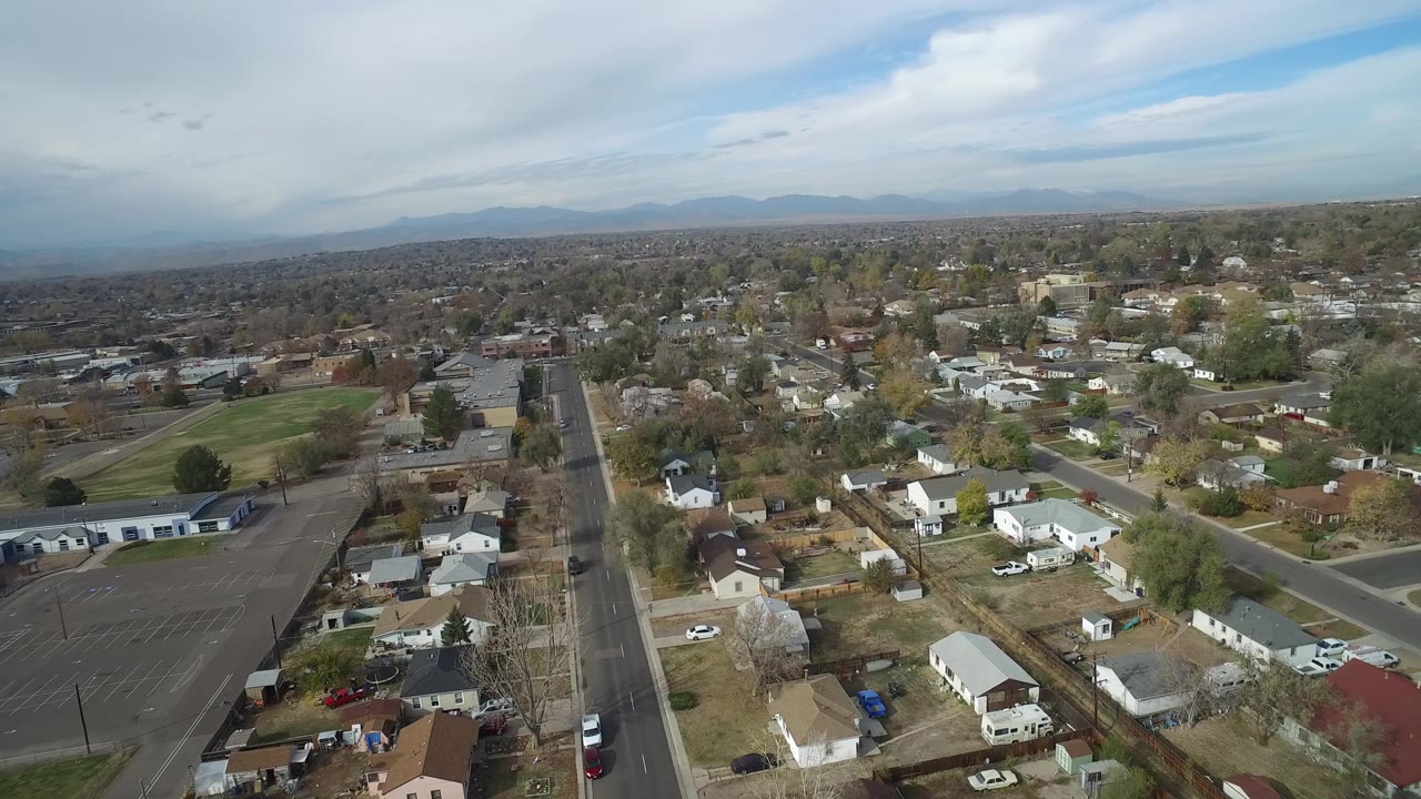 Aerial view over Hidden Lake, Colorado, with a straight neighborhood road at left, a large parking lot at lower right, low homes beyond, distant mountains, and no visible people.