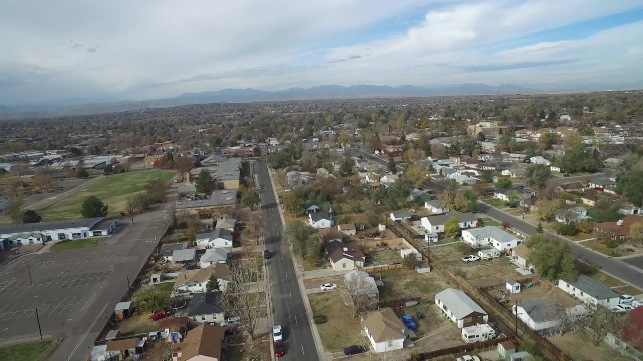 Wide aerial view of a residential area with a road, parking lot, homes, trees, and mountains.
