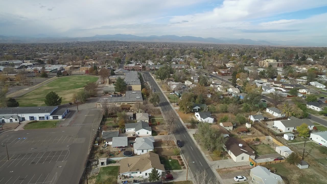 Elevated view across Hidden Lake, Colorado rooftops and parking spaces beside a straight road, with a grass field to the left and no clearly visible people.