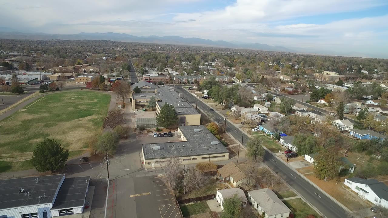 High aerial view in Hidden Lake, Colorado showing a large grassy school field beside flat-roofed buildings, neighborhood streets, homes, trees, distant mountains, and no visible people.