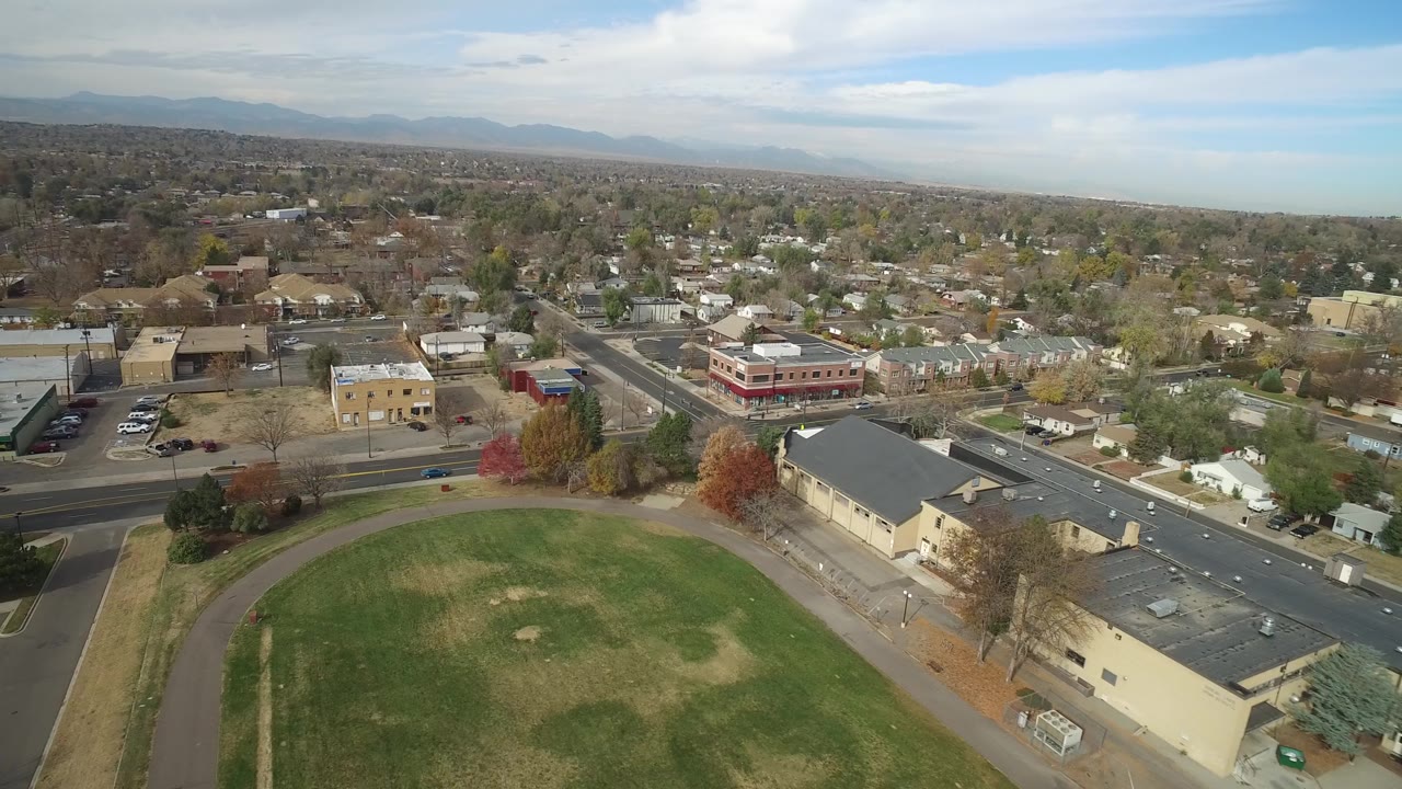 Aerial view in Hidden Lake, Colorado, looking across a green field toward intersecting roads, tan and red buildings, scattered trees, houses, distant mountains, and no visible people.