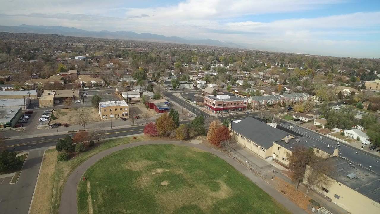 Drone-height view of town streets, buildings, trees, and a grass field.