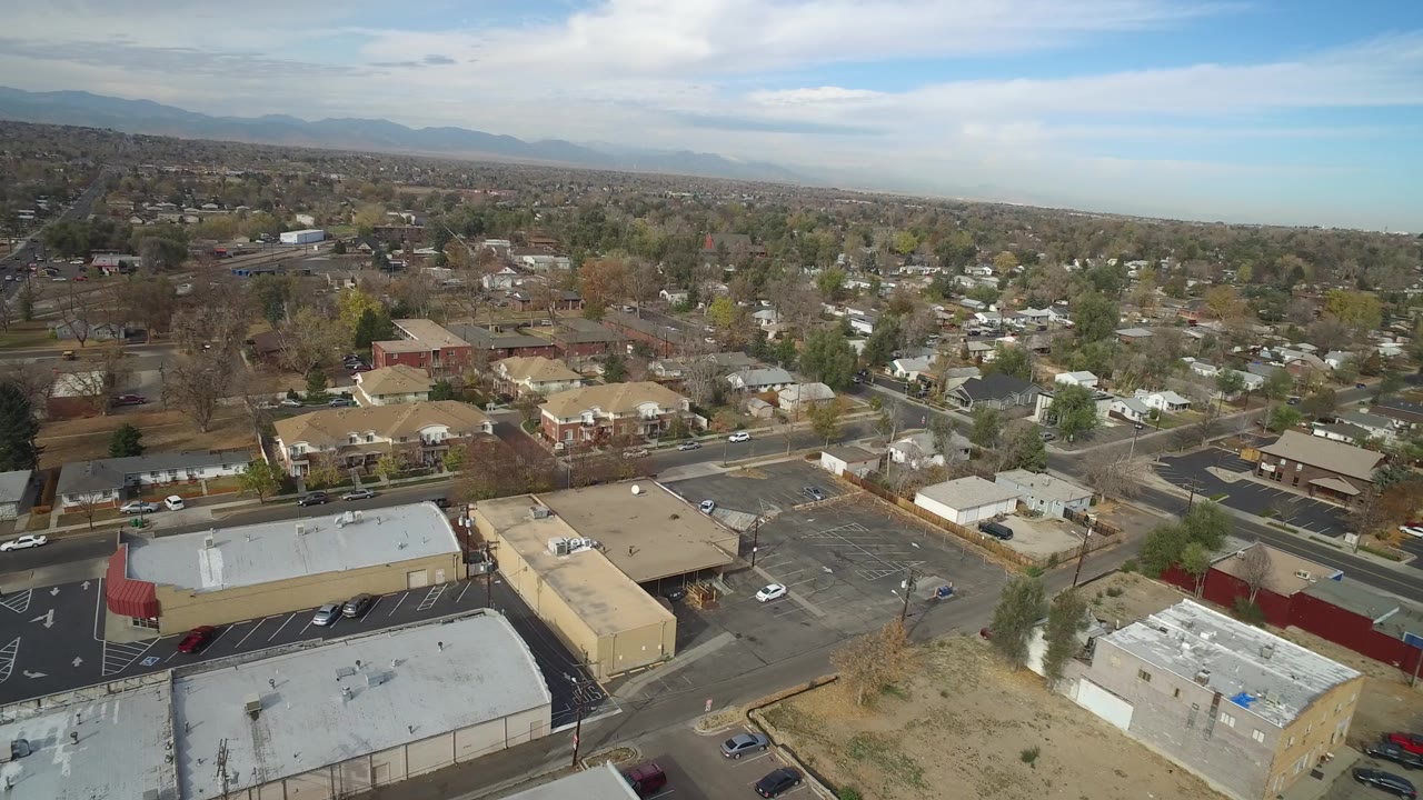 Opening aerial frame over Hidden Lake, Colorado, showing low commercial buildings, parking lots, residential blocks, trees, distant mountains, and no visible people.