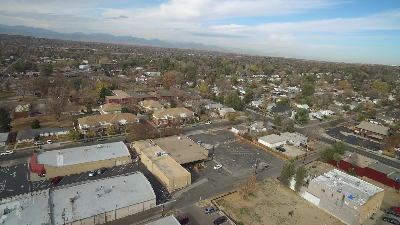 Aerial view in Hidden Lake, Colorado with a flat-roof commercial building in the foreground, nearby homes, parked cars, tree-lined streets, distant mountains, and no visible people.
