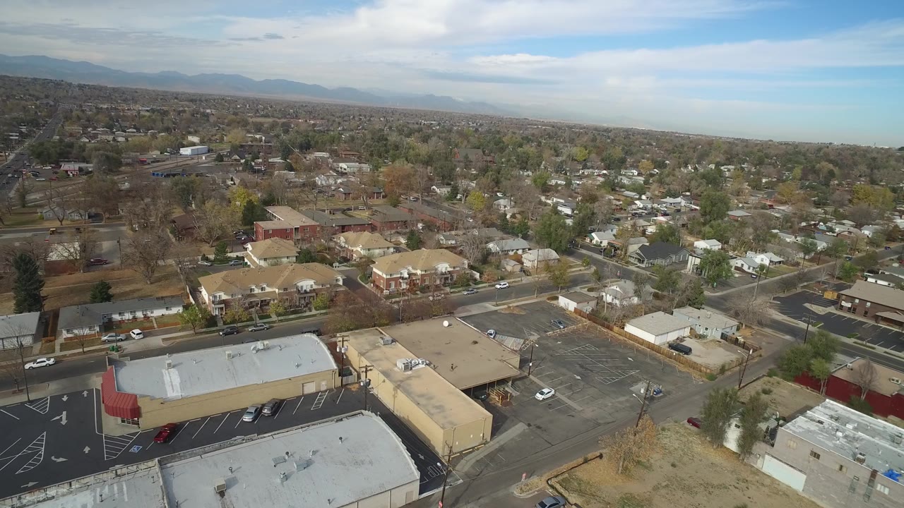 Aerial town frame with a large parking lot, tan buildings, houses, trees, and horizon.