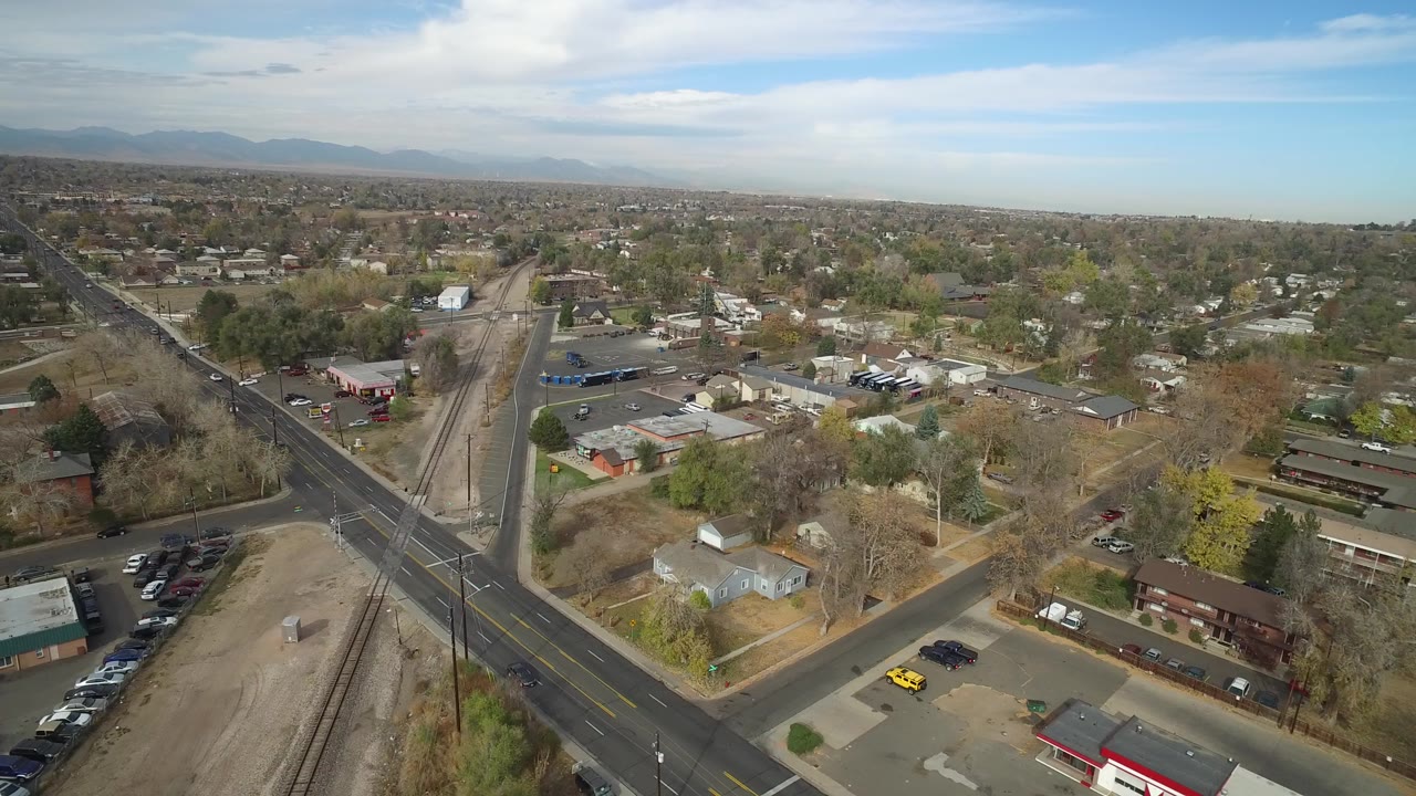 Wide aerial frame over Hidden Lake, Colorado, showing intersecting roads, a rail corridor, scattered homes, commercial buildings, distant mountains, blue sky, and no visible people.