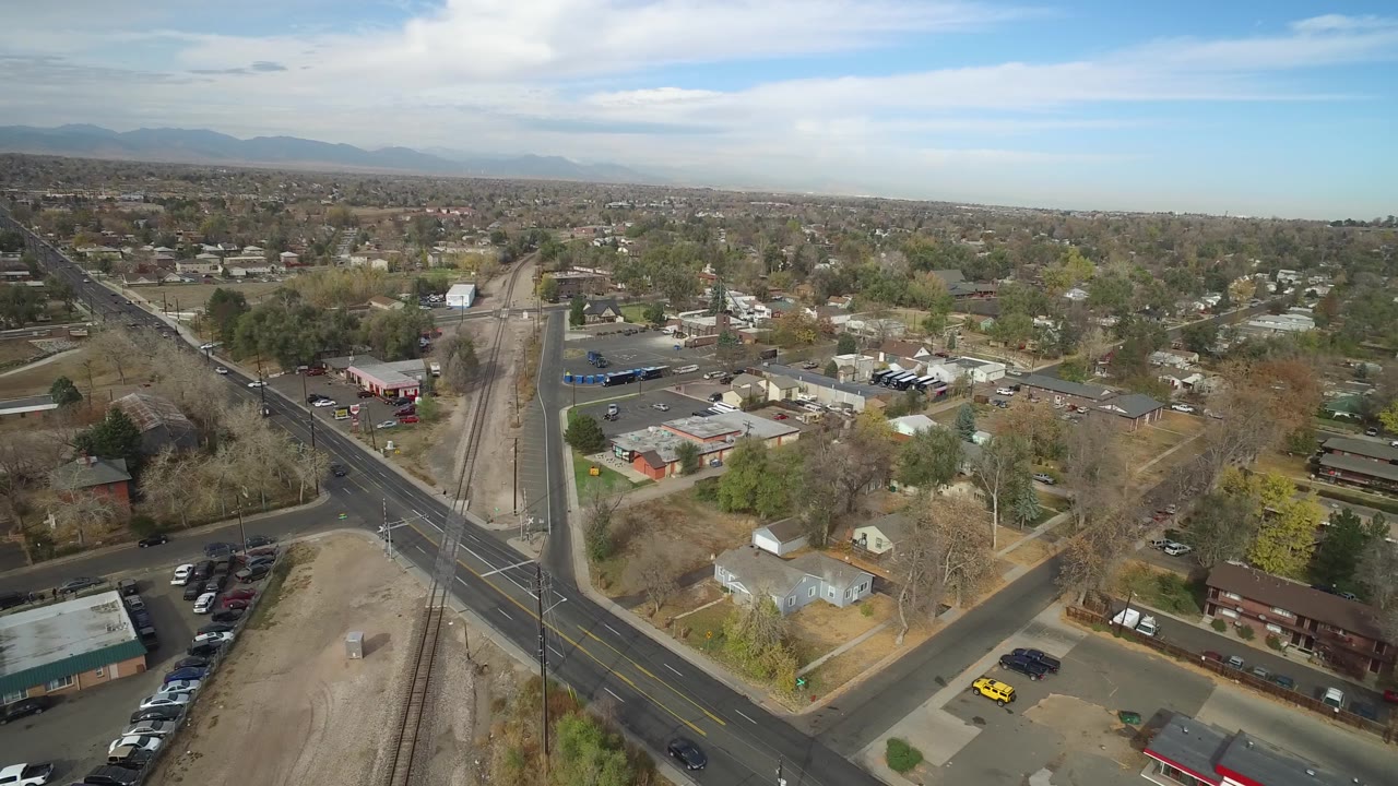 Drone-like aerial over a road crossing, rail corridor, homes, and parking lots.