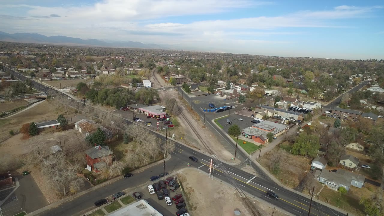Opening aerial frame over Hidden Lake, Colorado with intersecting roads, a small commercial strip, detached homes, brown grass, distant mountains, and no visible people.