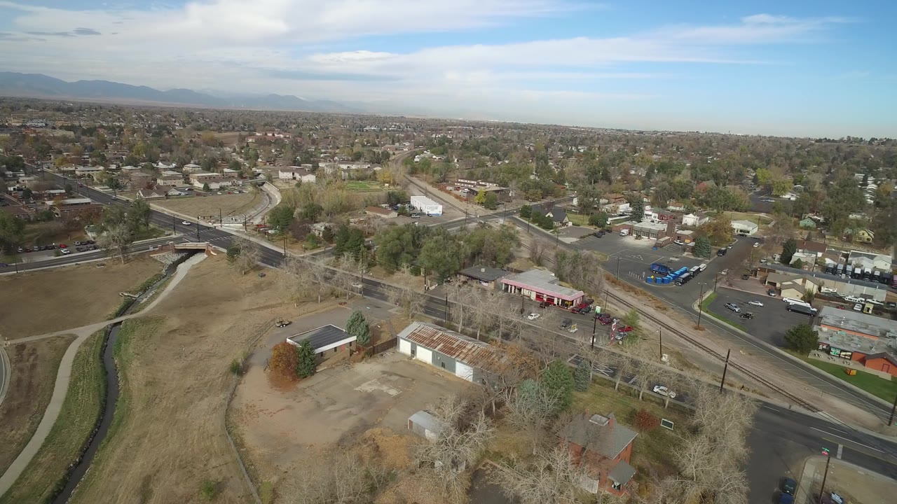 A wide drone view over Hidden Lake, Colorado shows a curving road, dry open ground, small buildings, residential blocks, distant mountains, and no visible people.