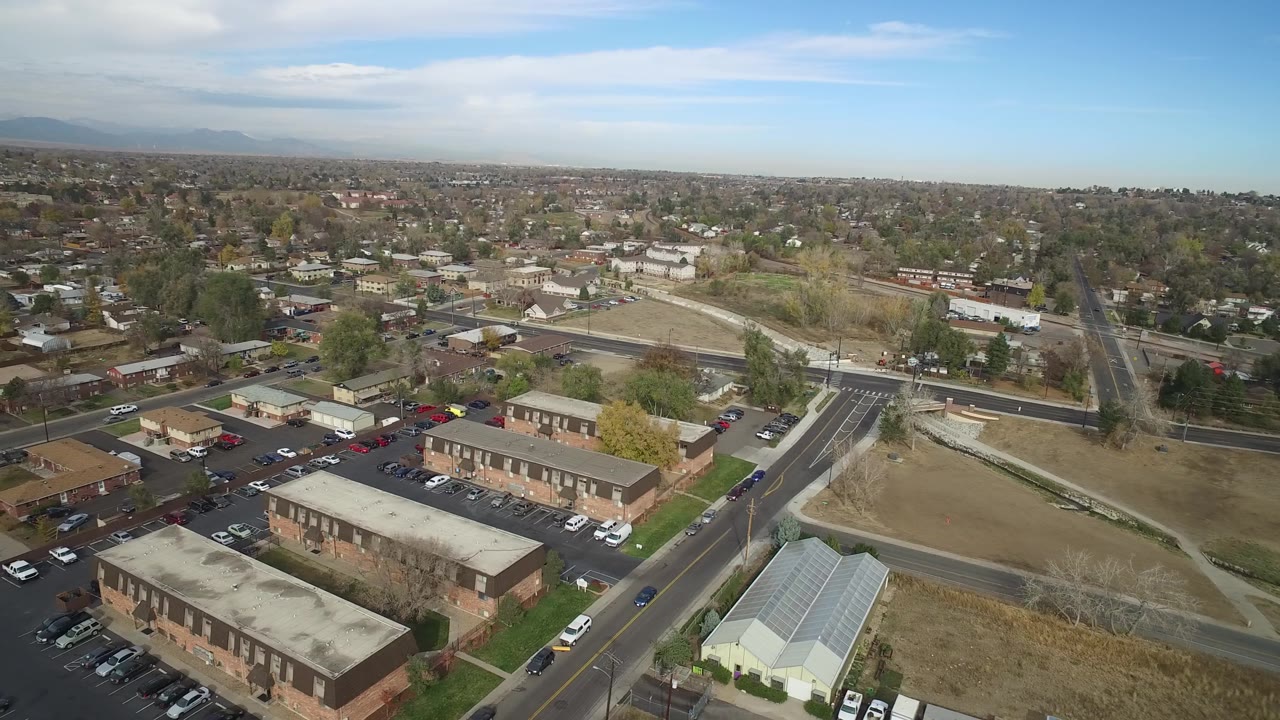 Aerial frame from Hidden Lake, Colorado, showing foreground parking lots and brick buildings, a pale open lot beside the road, clustered trees, distant residential blocks, and no visible people.