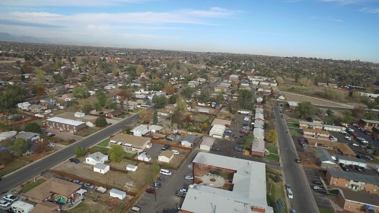 Aerial view of Hidden Lake, Colorado with low houses, a diagonal road, parked vehicles, open ground, tree cover, and a broad blue sky over the horizon.