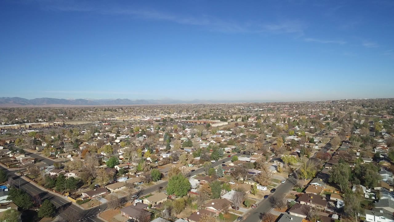 Wide aerial view over Hidden Lake, Colorado rooftops, streets, trees, distant mountains, and clear blue sky, with no visible people.