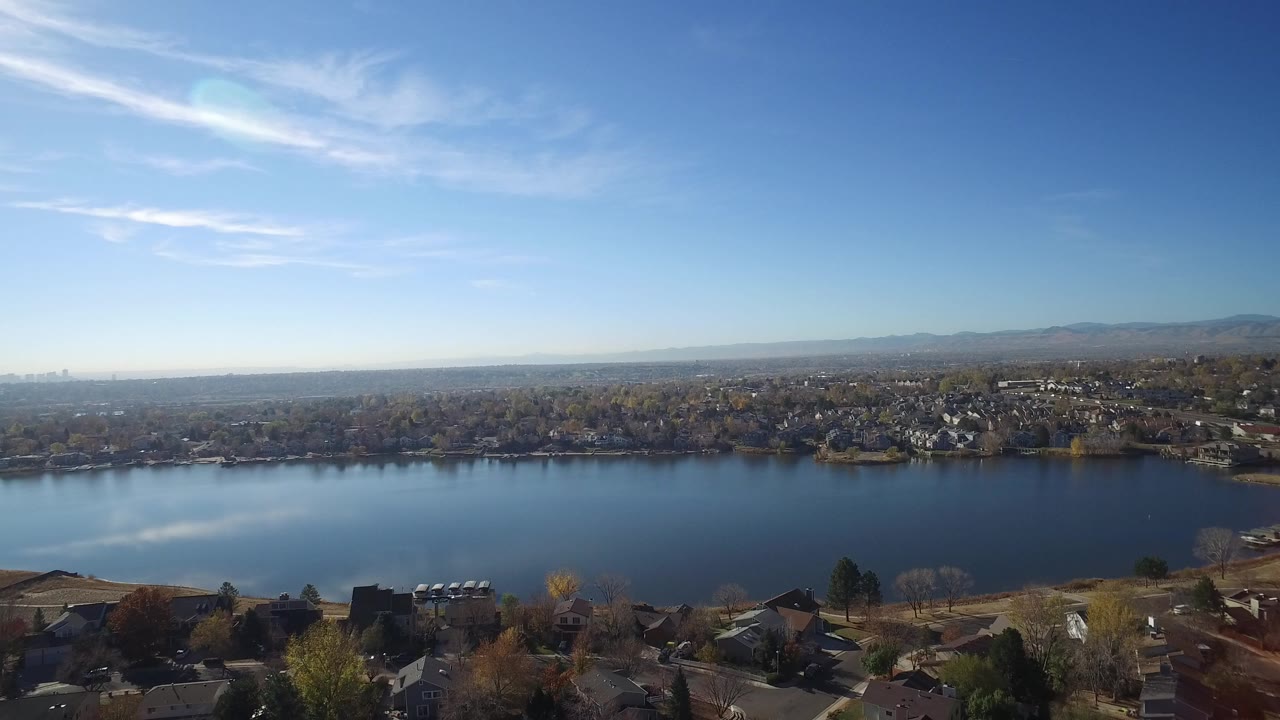 Aerial opening view of Hidden Lake, Colorado with blue water across the center, rooftops at the bottom edge, neighborhoods on the far shore, distant mountains, and no visible people.