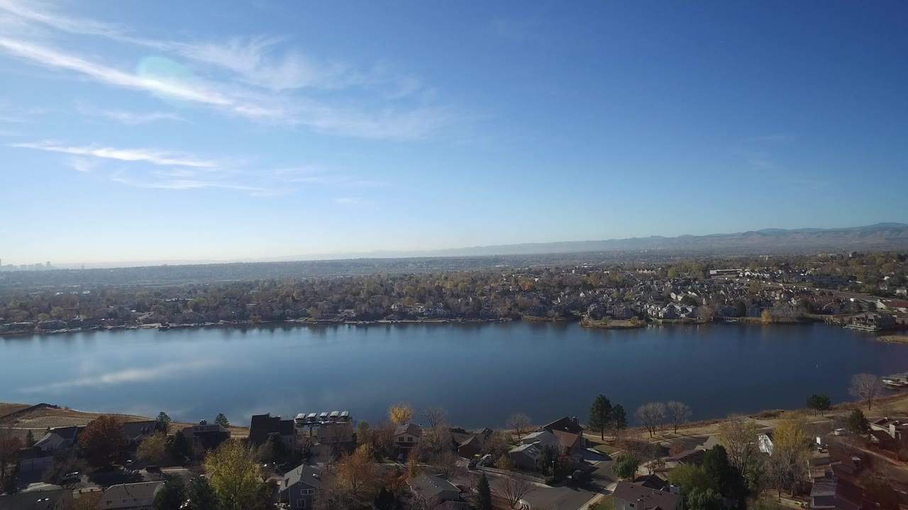 A high view over Hidden Lake, Colorado shows the lake stretching horizontally, suburban homes around the shoreline, a hazy mountain horizon, bright blue sky, and no visible people.