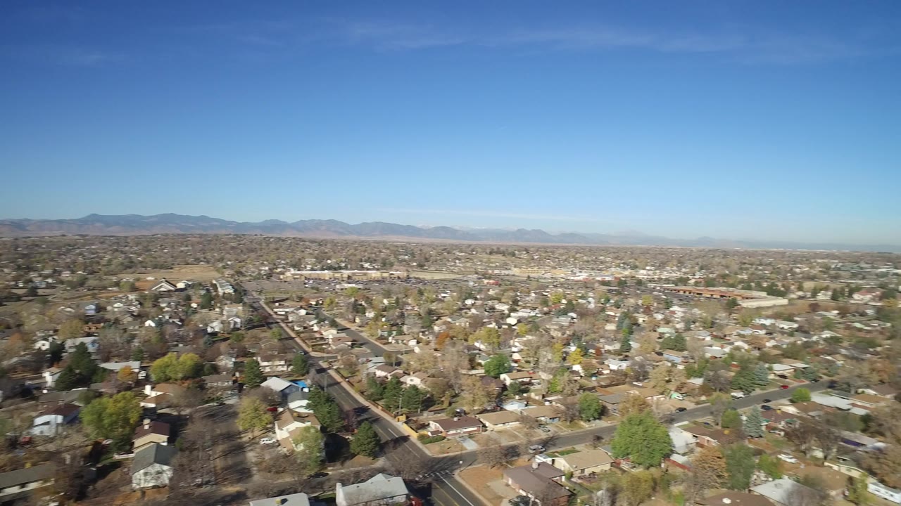 Aerial view across Hidden Lake, Colorado residential blocks, scattered trees, open lots, and low mountains on the horizon, with no visible people.