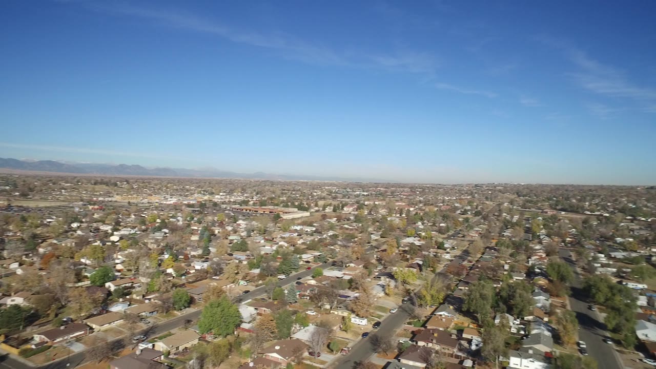 High aerial frame of Hidden Lake, Colorado showing dense residential blocks, tree-lined streets, tan ground areas, and a wide blue sky, with no visible people.