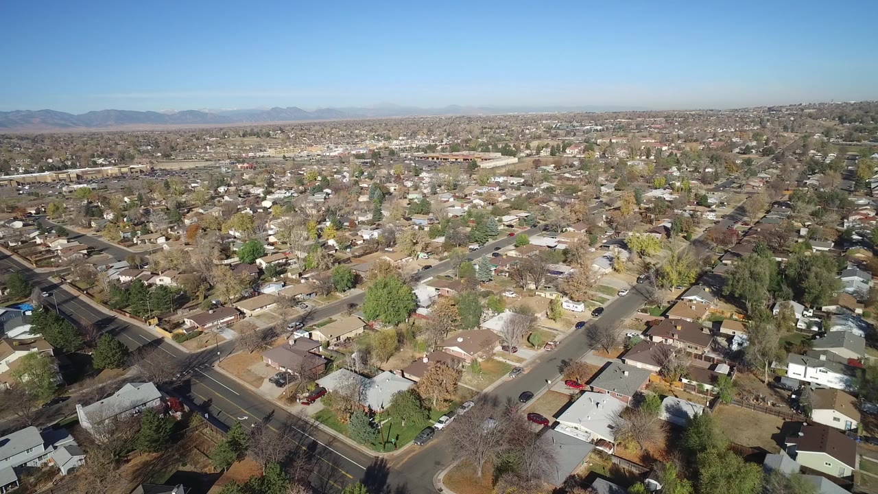 Aerial view over a suburban neighborhood with streets, houses, trees, and distant mountains.