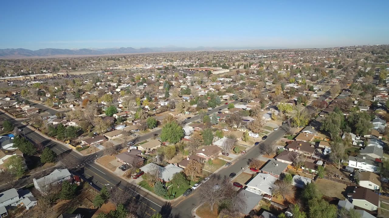 Aerial frame over Hidden Lake, Colorado, with a diagonal road crossing the lower view, compact homes and yards in the center, and mountains faintly visible on the horizon, with no visible people.