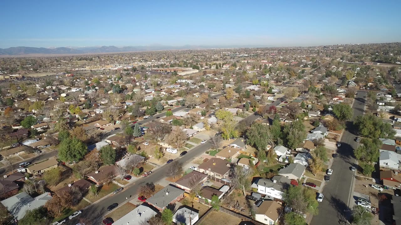 Aerial view over suburban houses and tree-lined streets under a clear sky.