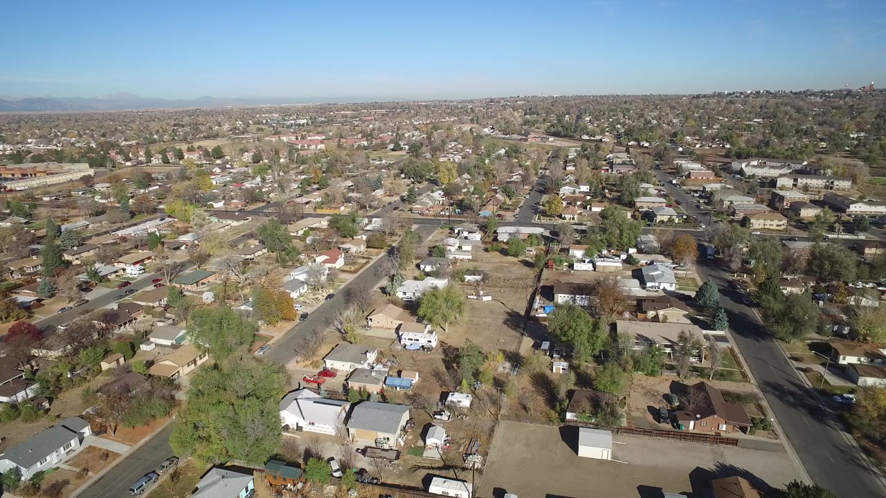 Aerial frame over Hidden Lake, Colorado, with a diagonal street at left, rooftops and dry yards below, distant residential blocks, and no visible people.