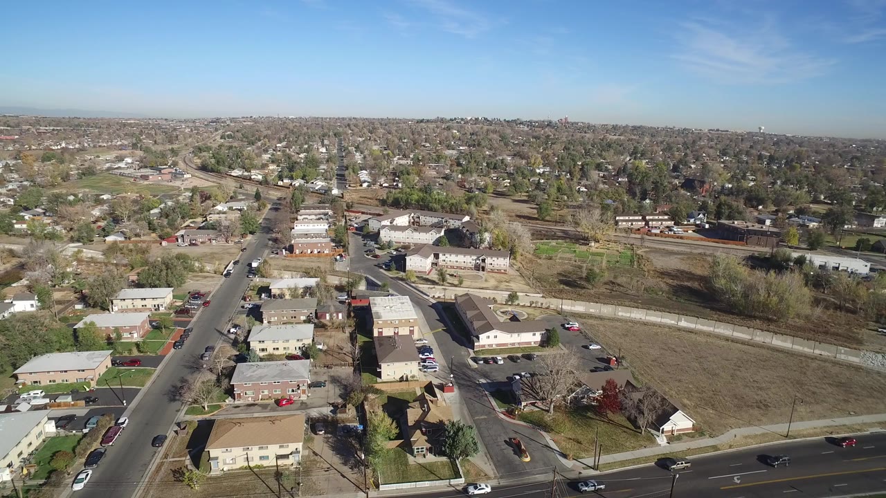 Aerial view of suburban homes, roads, open land, and a distant horizon under blue sky.