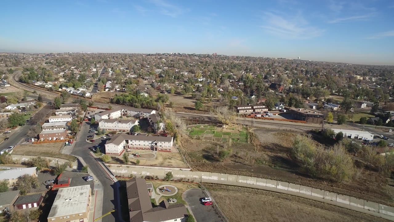 Wide overhead-oblique frame in Hidden Lake, Colorado showing low-rise residential buildings, a curving road, fenced open land, scattered trees, parked vehicles, and no visible people.
