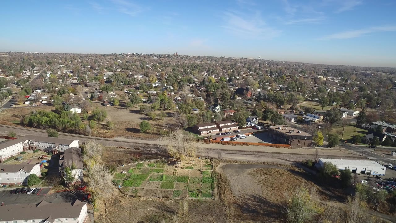 Aerial view over townhomes, garden plots, a road, trees, and distant houses.