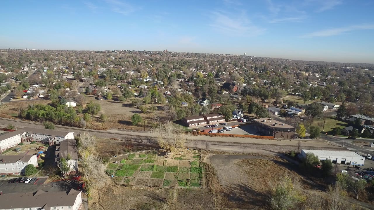 Aerial view in Hidden Lake, Colorado, with the road crossing the lower middle, brown open land and garden beds below it, townhomes and tree-lined houses beyond, and no visible people.