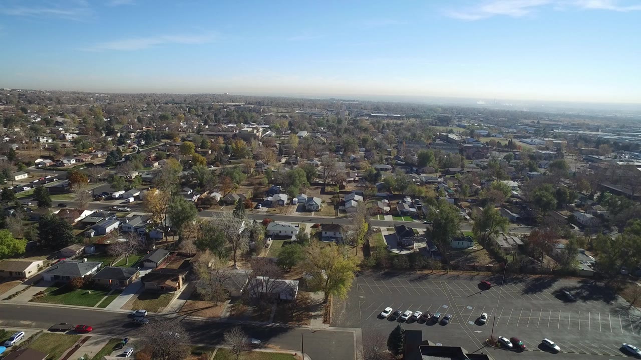 High aerial view of Hidden Lake, Colorado with detached houses, tree-lined residential streets, a parking lot at the bottom edge, and a wide blue sky above the horizon.