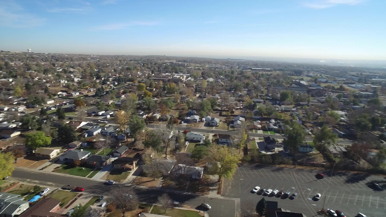 Aerial view over a residential subdivision with trees, streets, rooftops, and blue sky.