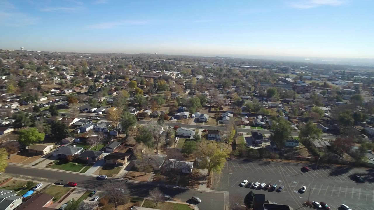 At 0:01, the Hidden Lake aerial remains over a leafy residential area, with houses and lawns below, a parking lot near the bottom edge, distant development on the horizon, and no visible people.