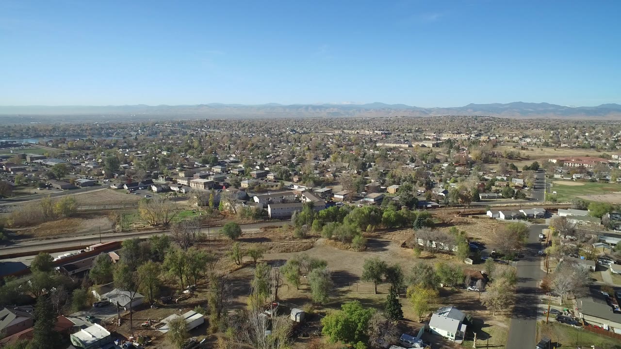 Opening aerial frame over Hidden Lake, Colorado, showing neighborhood streets, scattered houses, open lots, a bridge or rail crossing, distant mountains, blue sky, and no visible people.
