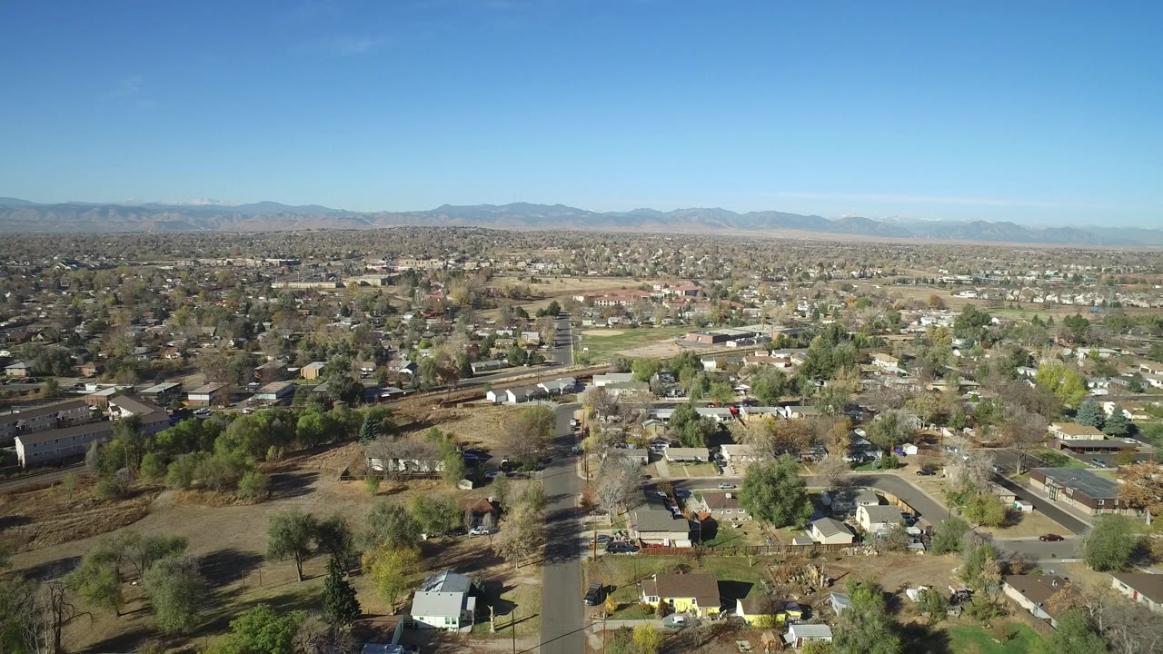 Wide aerial view over homes, roads, trees, and distant mountains under blue sky.