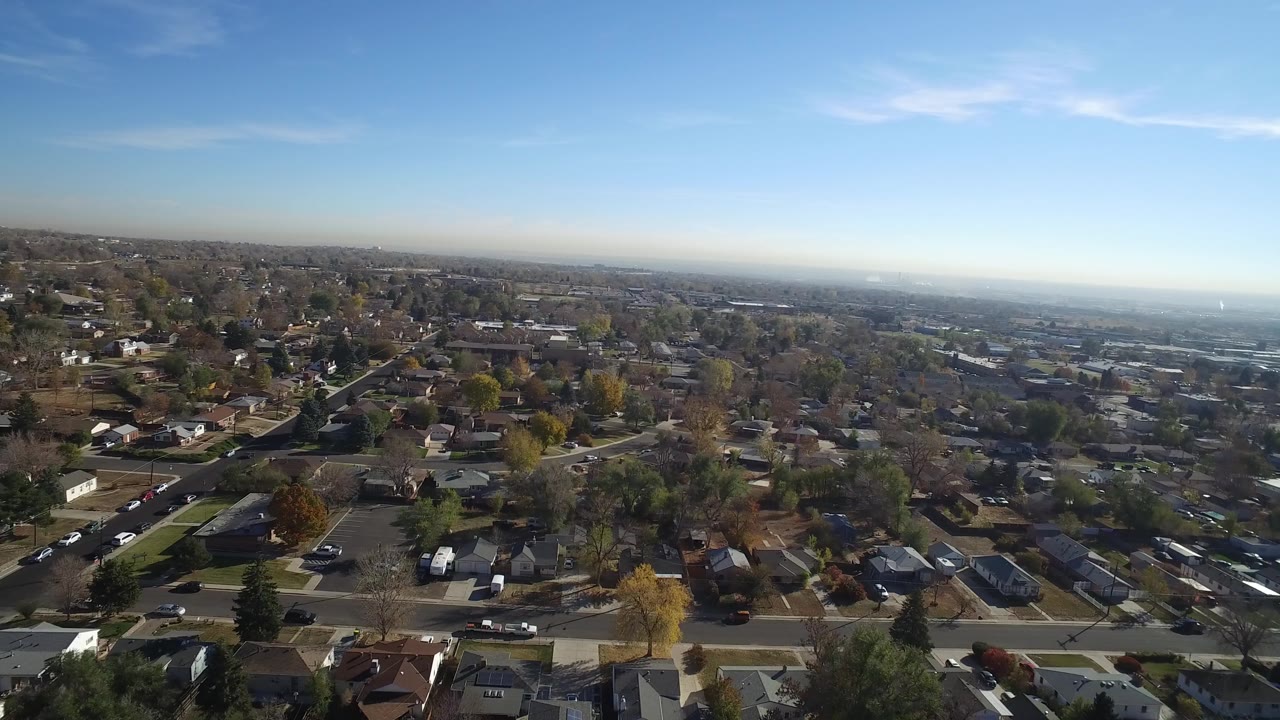 Aerial view over houses, streets, trees, and parked cars under blue sky.