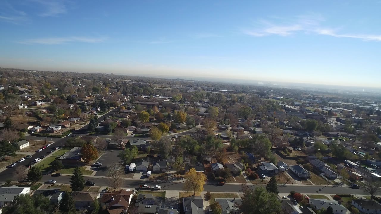 Early aerial frame in Hidden Lake, Colorado, with rooftops and streets in the foreground, scattered trees through the midground, distant buildings, and no visible people.