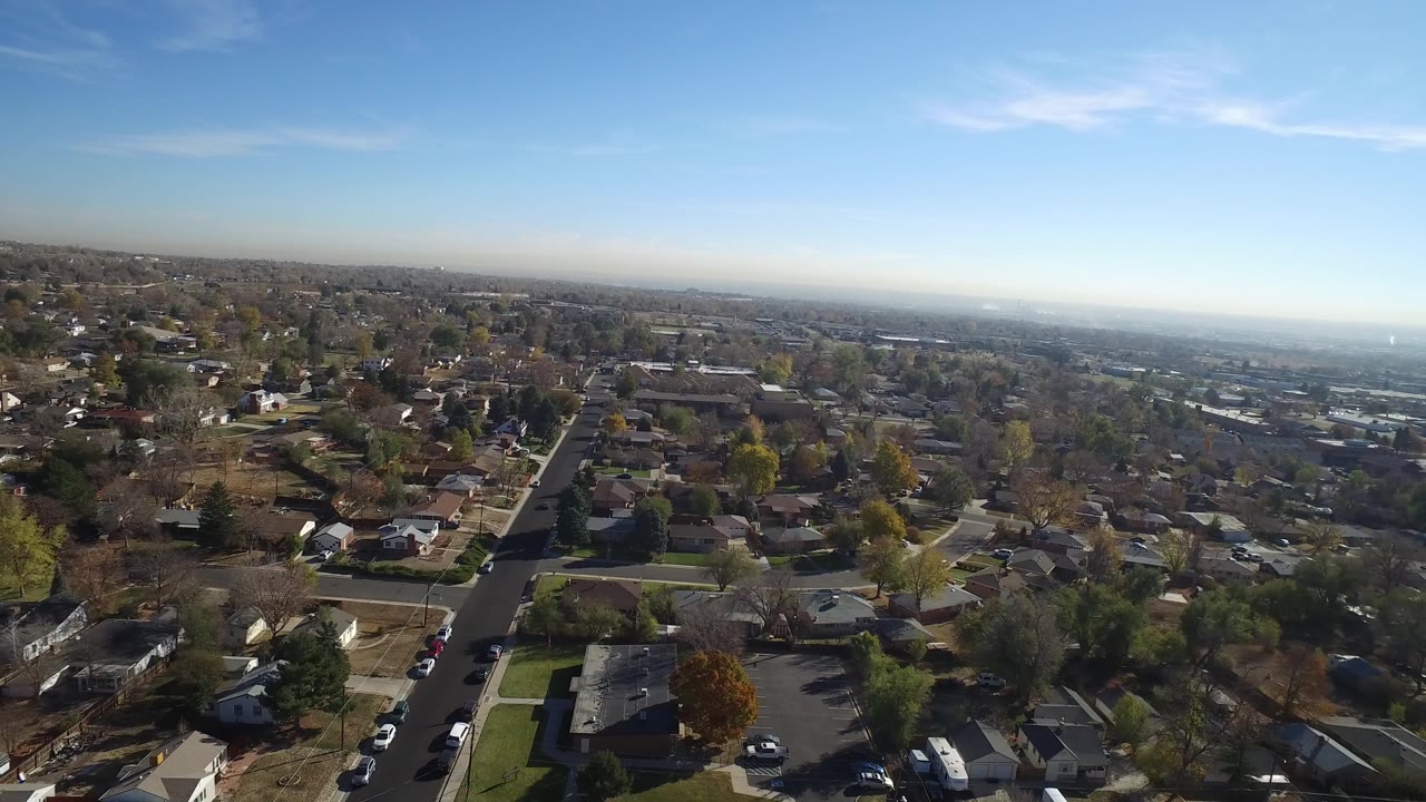 Opening aerial frame over Hidden Lake, Colorado, showing a residential street running toward houses and trees, with distant development on the horizon and no visible people.