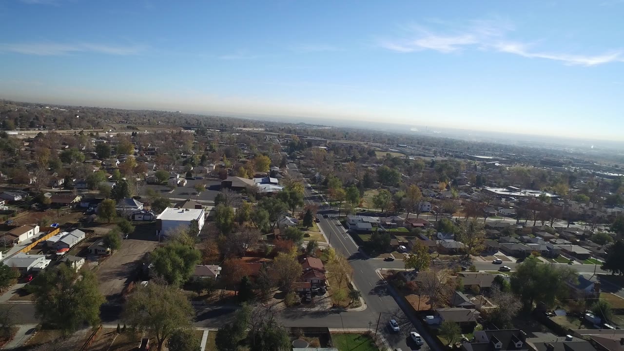 Suburban aerial with rooftops, intersecting streets, cars, trees, and open sky.