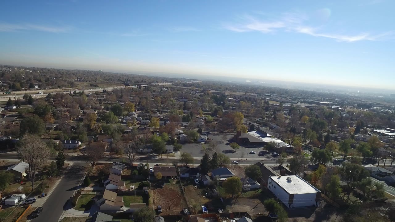 Opening aerial view over Hidden Lake, Colorado, with highway traffic at left, houses and trees below, a hazy horizon in the distance, and no visible people.