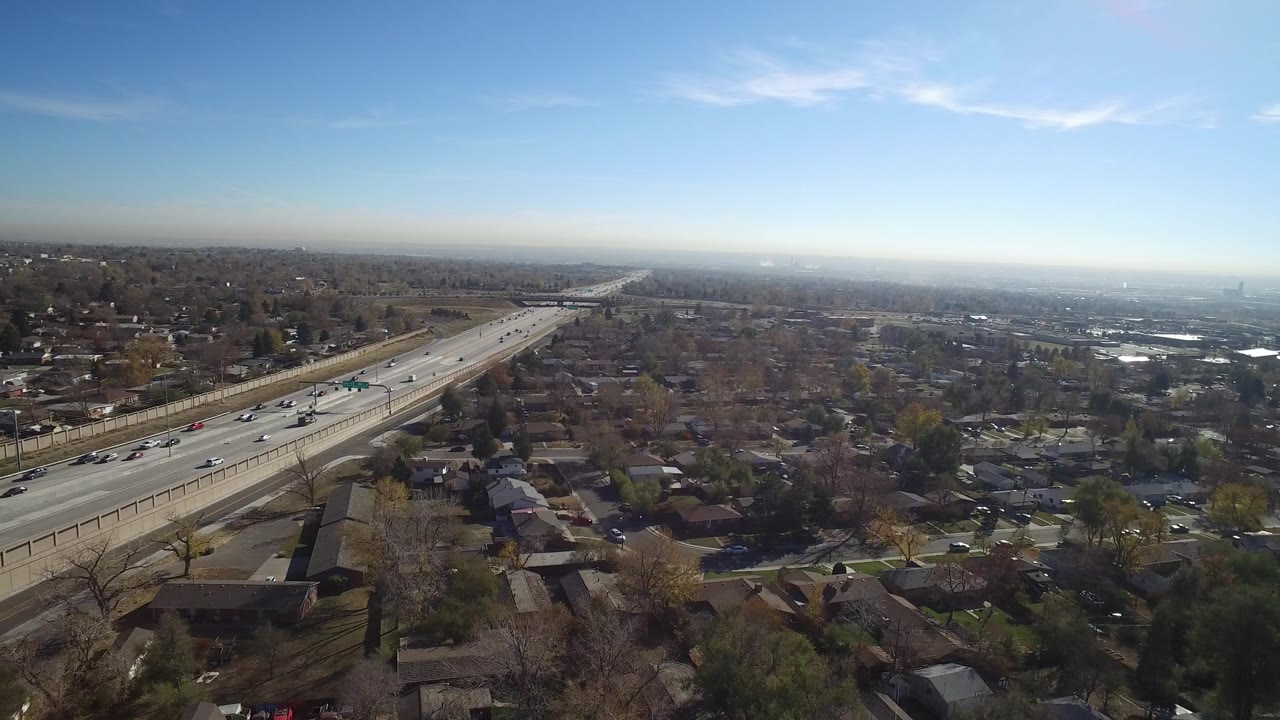 Aerial view in Hidden Lake, Colorado shows cars spread across highway lanes, residential blocks on both sides, bare trees, and a bright blue sky with no visible people.