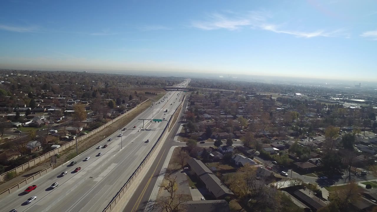 Opening aerial frame over Hidden Lake, Colorado shows a multi-lane road with moving cars, homes on both sides, a hazy horizon, and no visible people.