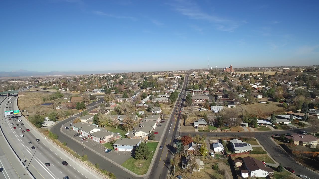 Aerial frame of Hidden Lake, Colorado with highway lanes on the left edge, small houses and intersecting neighborhood roads in the center, open fields beyond, and no visible people.