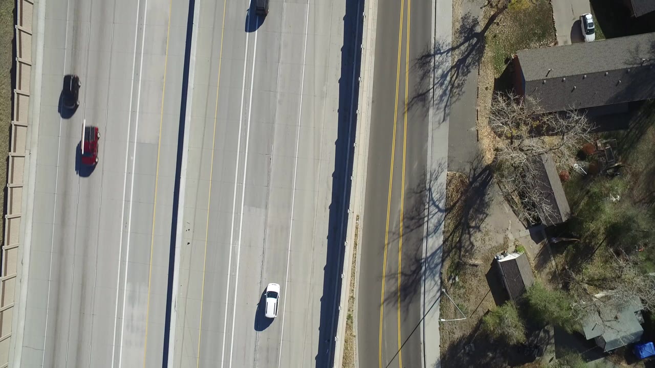 Overhead view of a multi-lane highway in Hidden Lake, Colorado, with a white car, darker vehicles, nearby rooftops, yards, and no visible people.