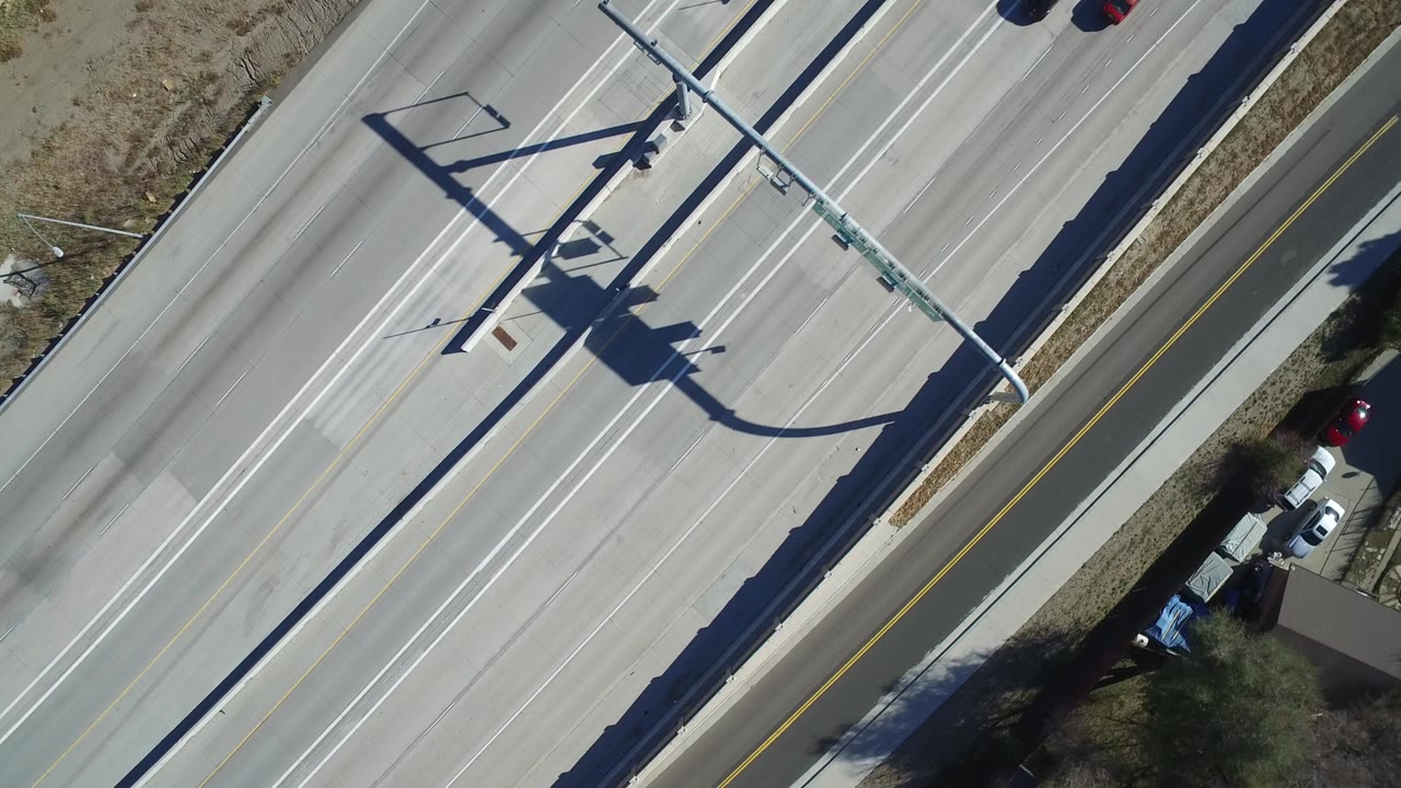 Aerial top-down view of a multi-lane highway in Hidden Lake, Colorado, with bright midday pavement, sign gantry shadows, roadside grass, and no visible people.