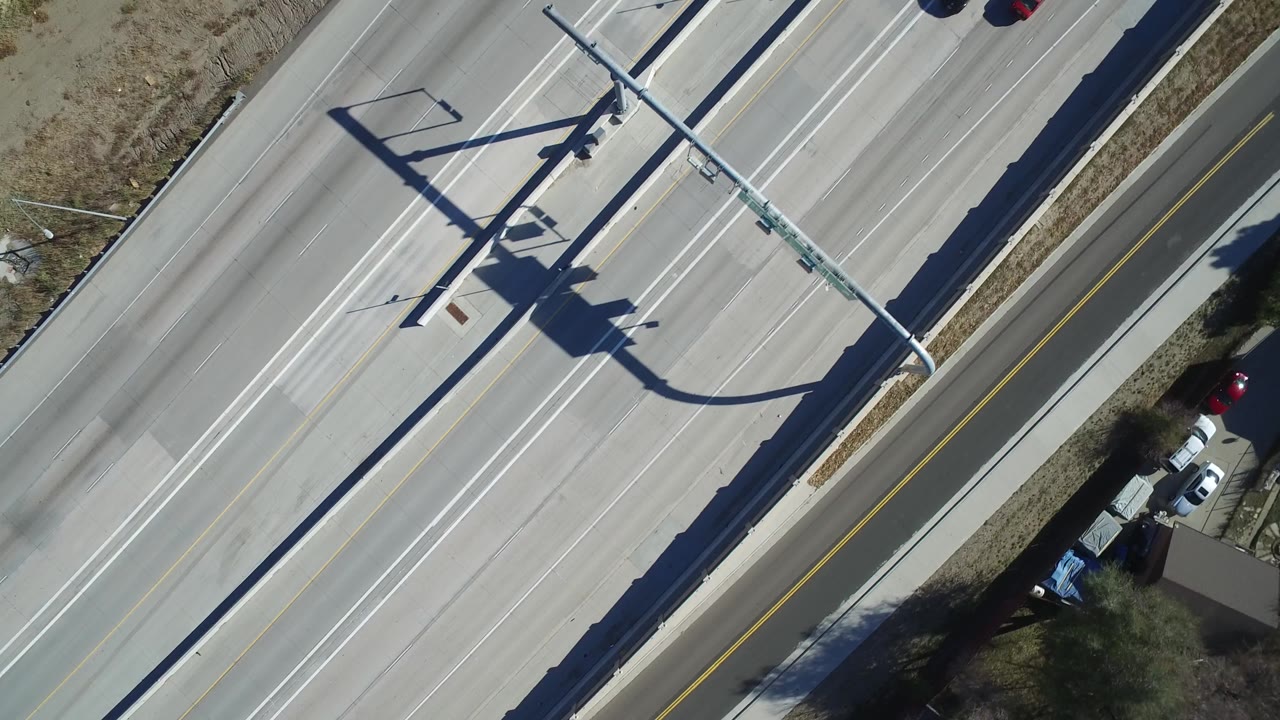 Top-down highway frame in Hidden Lake, Colorado, showing empty lanes, an overhead sign structure, a frontage road, parked vehicles near a home edge, and no visible people.
