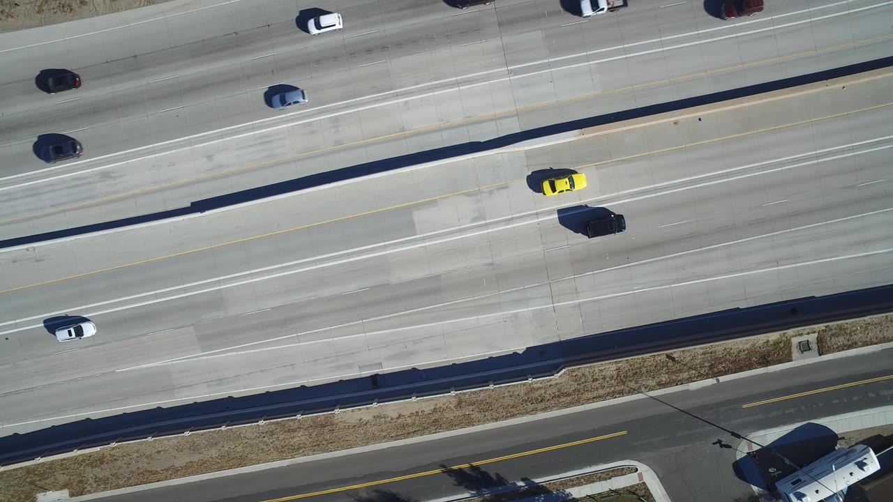 Overhead view of a sunlit divided highway in Hidden Lake, Colorado, with several cars, a yellow vehicle near the center lanes, roadside grass, and no visible people.