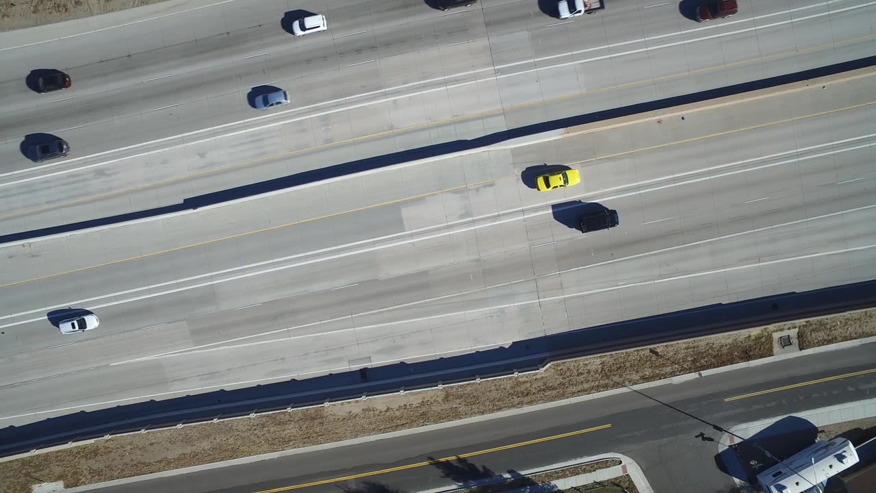 Top-down Hidden Lake, Colorado highway frame with the yellow car farther along the center lanes, a dark truck nearby, scattered vehicles, and no visible people.