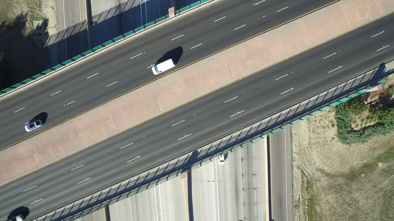 Overhead frame from Hidden Lake, Colorado showing a multi-lane highway, diagonal overpass edge, roadside grass, and several vehicles in bright sun with no visible people.