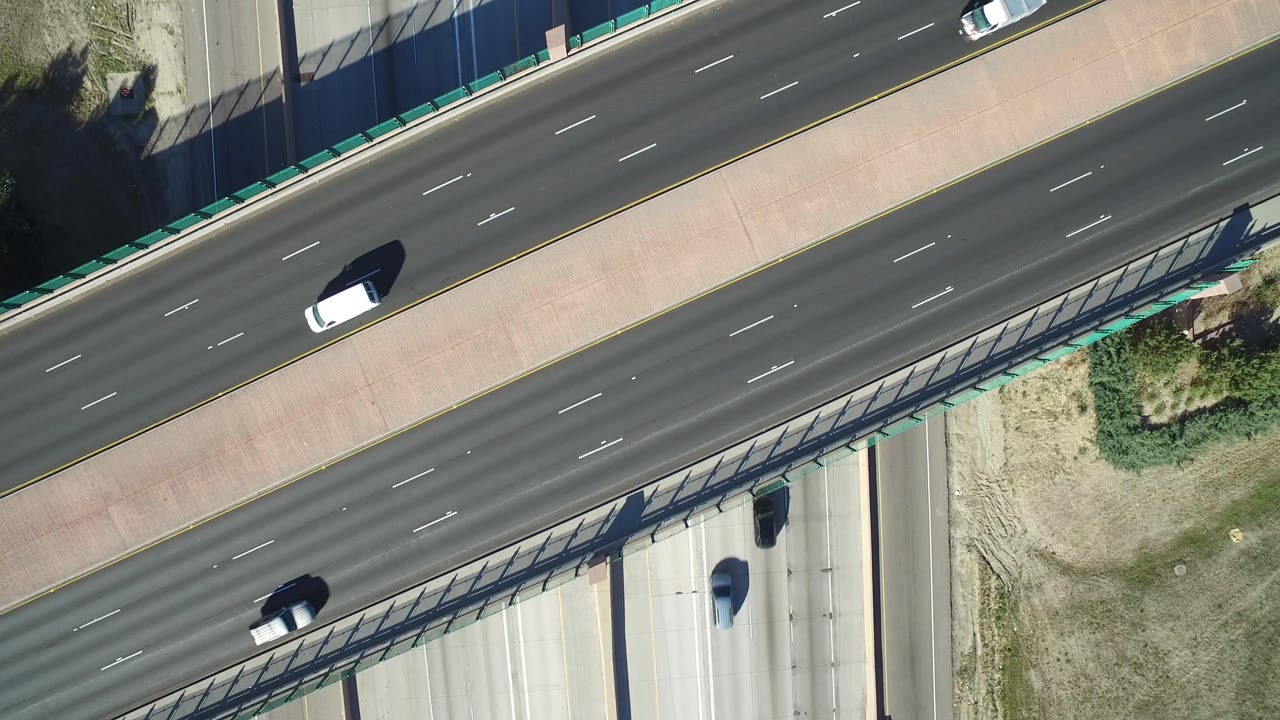 High overhead Hidden Lake, Colorado frame showing sparse cars on dark asphalt lanes, a concrete overpass strip, green guardrail detail, and shadow patterns with no visible people.