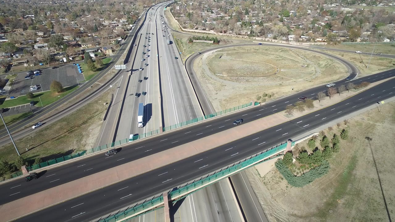 Hidden Lake, Colorado aerial view with cars and a white truck on the highway, a wide overpass crossing the frame, pale open ground, curved ramps, distant homes, and no visible people.